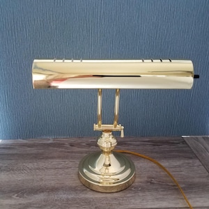 May include: A polished brass piano lamp featuring a long, cylindrical horizontal shade with a series of narrow ventilation slots along the top. The lamp has an adjustable arm connected to a round, weighted base and includes a thin brown power cord.