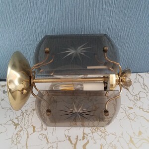 May include: Vintage brass wall sconce with two smoky glass shades. Each shade features a starburst design. The sconce has a gold finish and a decorative floral accent. The light fixture is mounted on a wall.