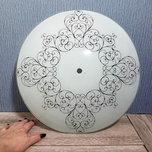 May include: A white, round ceiling light fixture with a black, ornate, symmetrical design. The design features swirling lines and heart-shaped elements. The fixture has a small hole in the center.
