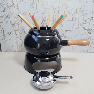 May include: A black fondue pot with a wooden handle and a stand. The pot has wooden fondue forks with colored tips. A silver burner is in front of the pot. The fondue pot is a classic design for entertaining.