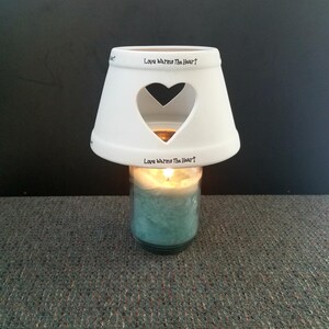 May include: A lit teal candle in a glass jar, topped with a white ceramic shade. The shade has a heart-shaped cutout and the text "Love Warms The Heart". The candle sits on a gray surface.