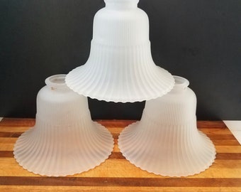 May include: Three frosted glass lamp shades with a fluted design. The shades are bell-shaped with a wide, ruffled bottom and a narrower top. They are stacked on a wooden surface against a black background.