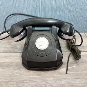 May include: A vintage black rotary phone with a handset and coiled cord. The phone has a circular dial with the text "ARIA705/RECON 567-3361 EXT 4". The handset has silver accents. The phone is on a wooden surface.