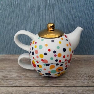 May include: A white ceramic teapot and teacup set with a gold lid and handle. The teapot and teacup are decorated with colorful polka dots in various sizes and colors, including red, yellow, blue, and pink. The set is designed for one person.