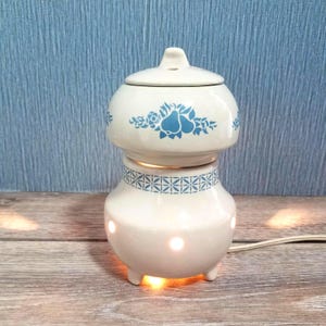 May include: A white ceramic oil warmer with a blue floral design and a lid. The warmer has a round base with three small feet and a patterned band. The top section has a blue floral design. The warmer is lit from within, and a power cord is visible.