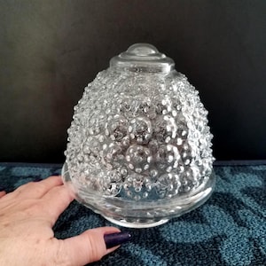 May include: Clear glass lamp shade with a textured, bubble-like surface. The shade has a rounded, bell-like shape with a small, circular top. The glass is transparent, allowing light to pass through. The shade is likely for a table lamp or ceiling fixture.