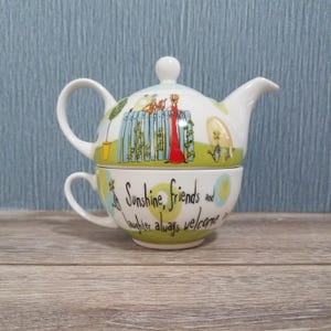 May include: A white ceramic teapot and teacup set. The teapot has a lid and spout, and the cup sits beneath it. The set is decorated with a whimsical design featuring a woman in a red dress and the text "Sunshine, friends and laughter always welcome."