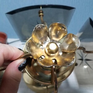 May include: Close-up of a vintage brass flower-shaped light fixture. The fixture has a gold finish with a central bulb holder and curved arms. The fixture is paired with a dark gray glass shade with a starburst design.