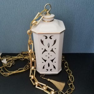 May include: White ceramic hanging lantern with gold trim and decorative cutouts. The lantern is suspended by a gold chain and has a matching tassel. The lantern is an octagonal shape.