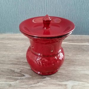 May include: A glossy, red ceramic jar with a matching lid. The jar has a bulbous base, a narrow neck, and a decorative band. The lid is topped with a small, pointed finial. The jar is likely for storage or decoration.