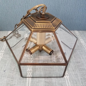 May include: Vintage brass and glass pendant light fixture with a geometric design. The fixture has a hexagonal shape with clear glass panels and a brass frame. It features three light bulb sockets and a chain for hanging.
