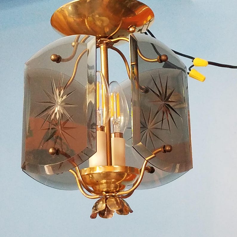 May include: Vintage brass ceiling light fixture with two light bulbs. The fixture has a gold finish and features etched glass panels with starburst designs. The light is suspended from a brass canopy and has a decorative floral base.
