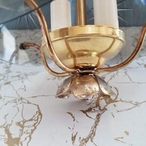May include: Close-up of a vintage chandelier with a gold-toned metal frame and a clear glass shade. The chandelier has three white candle-shaped lights and a decorative flower-shaped base. The fixture is set against a marble-patterned background.