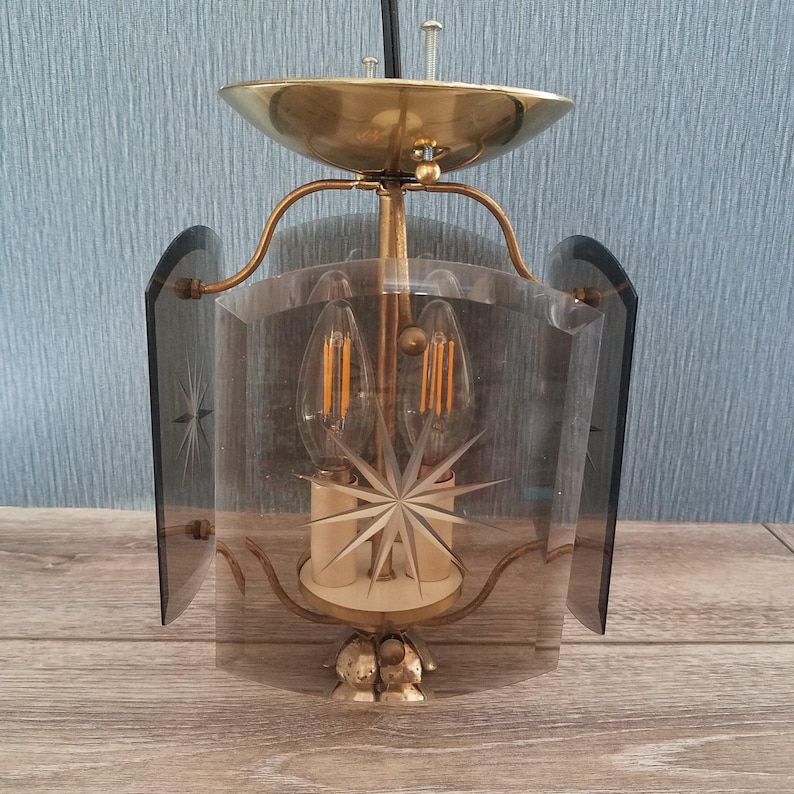May include: Vintage brass and glass pendant light fixture. The fixture features a brass canopy, arms, and base, with a clear glass shade etched with starburst designs. The light has two exposed bulbs and is suspended from a black cord.