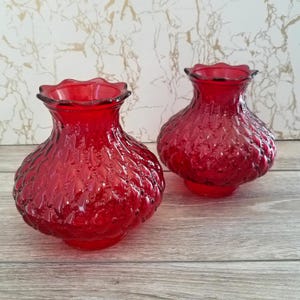 May include: Two red glass vases with a textured surface and a ruffled rim. The vases have a bulbous shape and sit on a small, round base. The background is a light, marbled pattern.