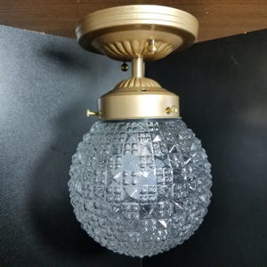 May include: A vintage ceiling light fixture with a gold-colored base and a clear, textured glass globe. The globe has a diamond-like pattern. The fixture is mounted on a wooden surface.