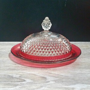 May include: A vintage glass butter dish with a ruby red base and a clear, diamond-patterned dome. The lid is topped with a decorative finial. The dish is designed to hold butter and keep it fresh.