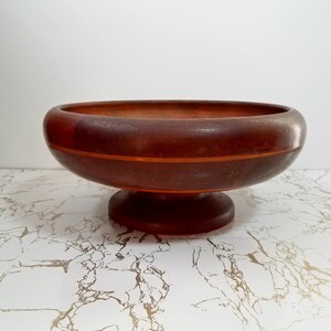May include: A dark brown wooden pedestal bowl with a wide, shallow design. The bowl features a circular base and a contrasting orange-toned stripe around the middle. The interior of the bowl is also dark brown.
