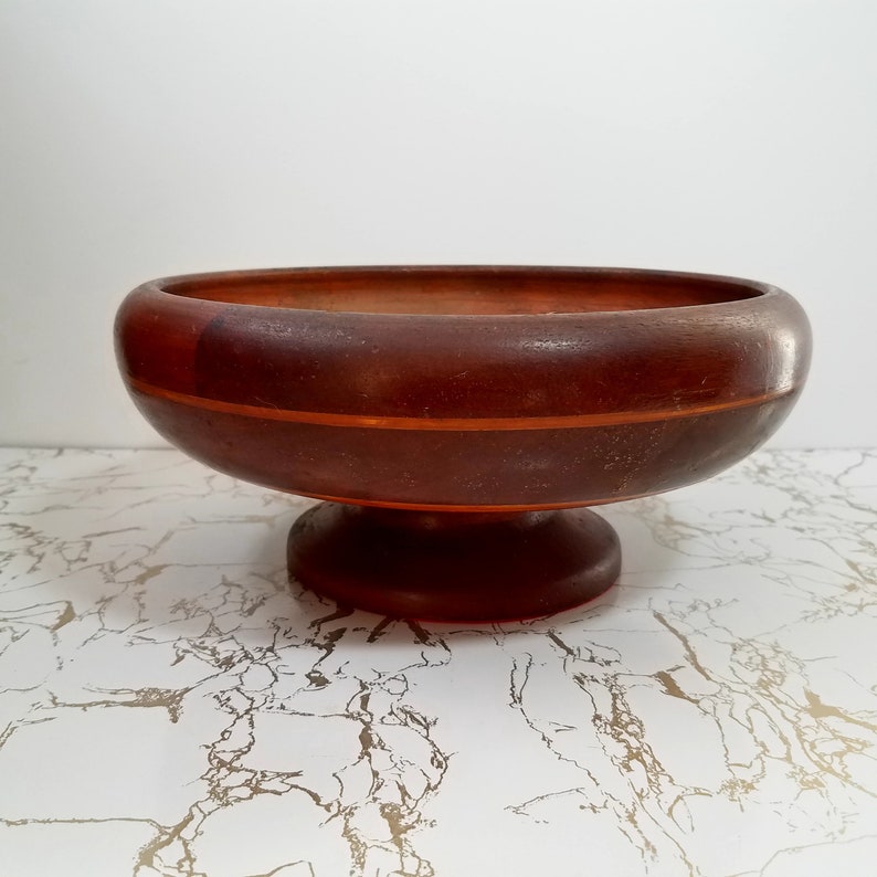 May include: A dark brown wooden pedestal bowl with a wide, shallow design. The bowl features a circular base and a contrasting orange-toned stripe around the middle. The interior of the bowl is also dark brown.