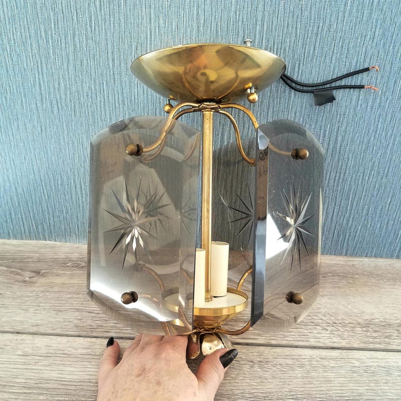 May include: Vintage brass and glass ceiling light fixture. The fixture features a brass canopy and decorative arms supporting a central column. The glass panels are clear with etched starburst designs. The fixture is designed to hold two light bulbs.