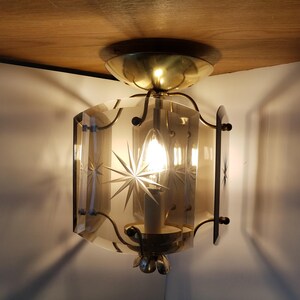 May include: A vintage ceiling light fixture with a starburst design etched into the glass panels. The fixture has a brass finish and a single light bulb. The light is on, casting shadows on the wall.