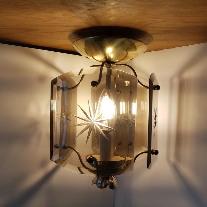 May include: A vintage ceiling light fixture with a starburst design etched into the glass panels. The fixture has a brass finish and a single light bulb. The light is on, casting shadows on the wall.