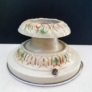 May include: Off-white vintage lamp base with decorative floral accents in green, pink, and brown. The base has a circular design with a central column and a small screw. The lamp base is made of metal.