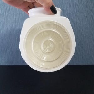May include: A white, hexagonal-shaped light fixture with a clear, circular, ridged glass lens. The fixture is held by a hand with a black painted fingernail. The background is a textured blue surface.
