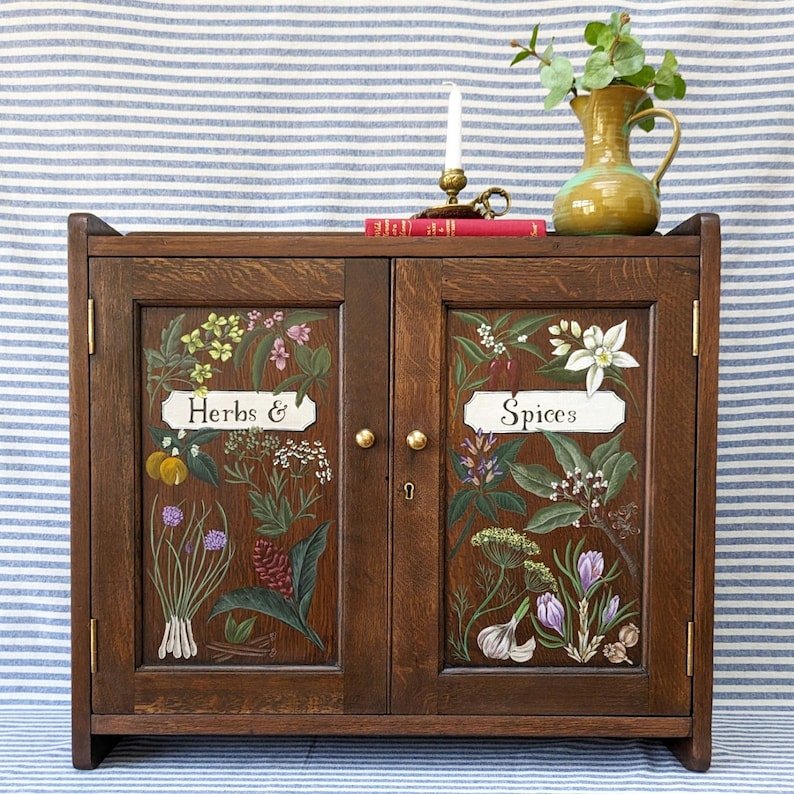 May include: A dark wood cabinet with two doors. The doors are painted with botanical illustrations of herbs and spices. The doors are labeled "Herbs &" and "Spices".