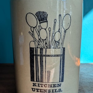 May include: A beige ceramic kitchen utensil holder with a black illustration of various kitchen tools. The holder has the words "KITCHEN UTENSILS." printed in black.