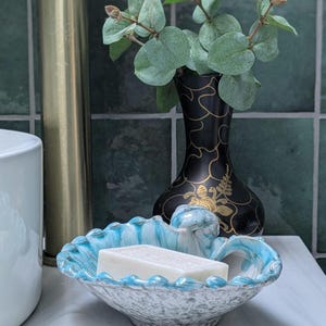 May include: A blue and white ceramic soap dish shaped like a seashell with a white bar of soap inside. The dish is sitting on a white countertop next to a black vase with green leaves.