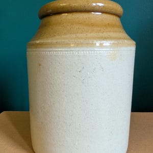 May include: A traditional stoneware jar, featuring a light beige body and a darker tan rim. The cylindrical jar has a slightly flared top and a decorative band. The background is a teal colour. The jar is likely used for storage.