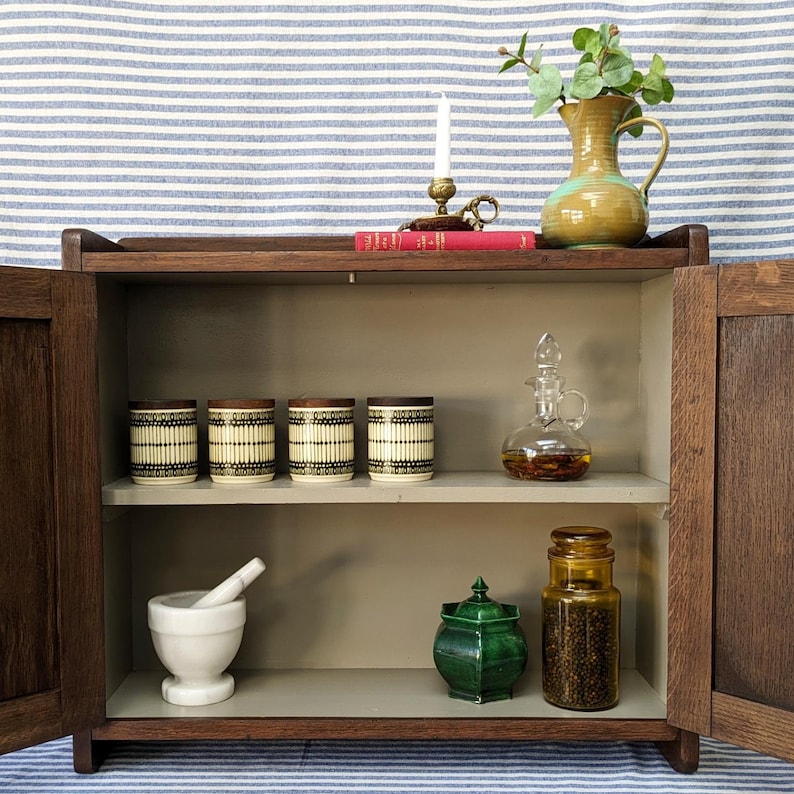May include: A wooden cabinet with two doors and two shelves. The top shelf has four small jars with black and white patterns, a red book, and a small candle holder. The bottom shelf has a white mortar and pestle, a green jar with a lid, and a clear glass bottle with a cork stopper.