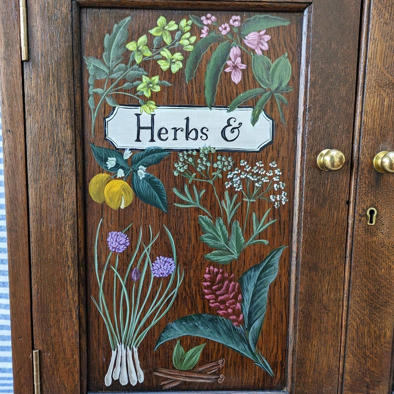 May include: A wooden cabinet door with a painted design of various herbs and flowers. The door is painted in a dark brown color and features a sign that reads "Herbs &" in black lettering.