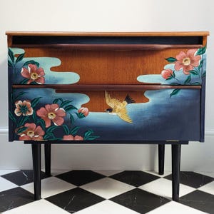 May include: A two-drawer wooden cabinet with a dark blue painted exterior. The cabinet features a floral and bird design in shades of pink, green, and yellow. The cabinet has black legs and a dark wood top.
