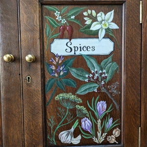 May include: A wooden cabinet door with a painted design of various spices and herbs. The door is painted in a dark brown wood grain pattern. The words "Spices" are painted in white on a white rectangular sign.