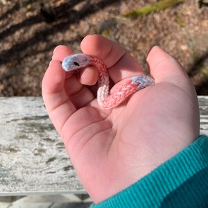 May include: A small, handmade crochet snake in shades of pink, white, and gray. The snake is held in a hand, showcasing its detailed stitching and design. The background is blurred, highlighting the craft item.