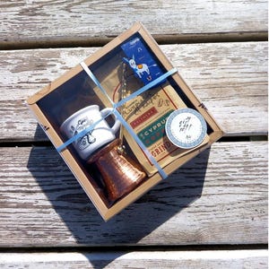 May include: A gift box set for making Greek coffee. The set includes a small white ceramic mug, a copper cezve, a bag of Cyprus coffee, and a donkey charm. The box is wrapped in brown paper with blue ribbon and a sticker that says "Gift Set!"
