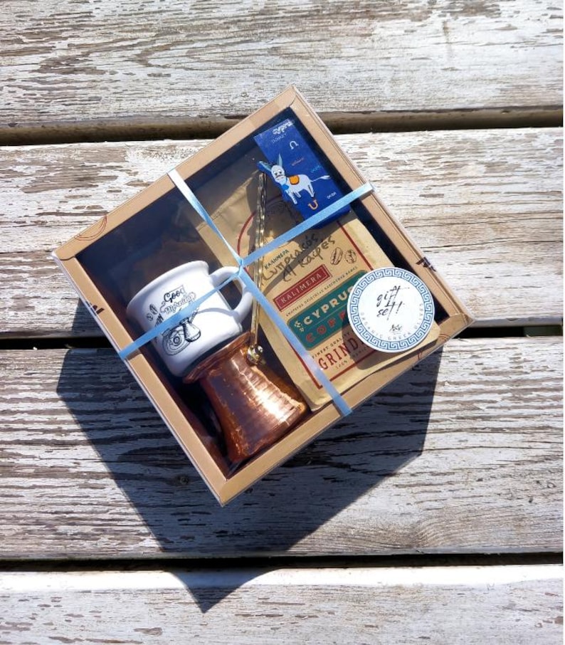 May include: A gift box set for making Greek coffee. The set includes a small white ceramic mug, a copper cezve, a bag of Cyprus coffee, and a donkey charm. The box is wrapped in brown paper with blue ribbon and a sticker that says "Gift Set!"