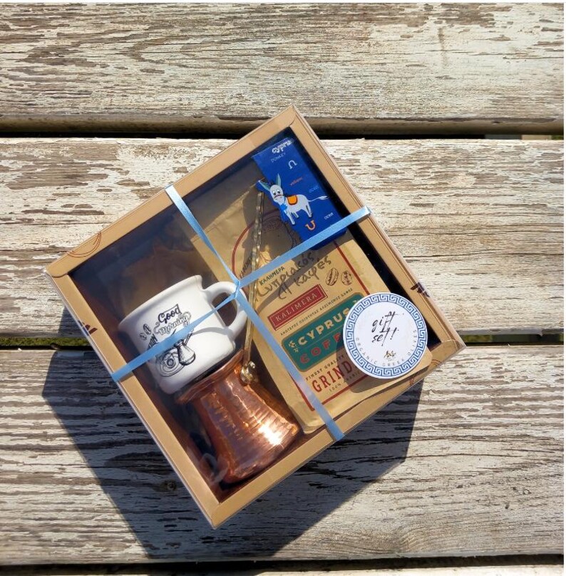 May include: A gift set for coffee lovers featuring a copper coffee pot, a white ceramic mug with the text "Good Morning" and a bag of Cyprus coffee. The set is packaged in a brown box with a blue ribbon and a sticker that says "Gift Set!!".