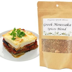 Op de afbeelding: Een stuk Griekse moussaka op een wit bord, gegarneerd met peterselie. Daarnaast een bruine papieren zak met "Greek Moussaka Spices Blend" met een transparant venster dat de kruidenmix laat zien. Het etiket luidt "Organic Greek Olives".