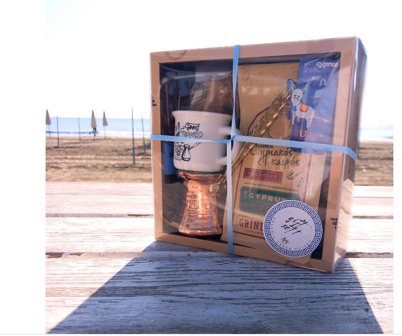 May include: A gift box set containing a white ceramic coffee cup with a Greek design, a copper coffee pot, and a bag of Greek coffee. The box is decorated with a blue and white Greek key design and the text "Cyprian Coffee".