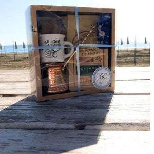 May include: A gift box set containing a small white ceramic coffee cup with a brown handle, a copper Turkish coffee pot, a bag of coffee grounds, and a blue and white sticker with a donkey illustration. The sticker reads "Cyprus Coffee".
