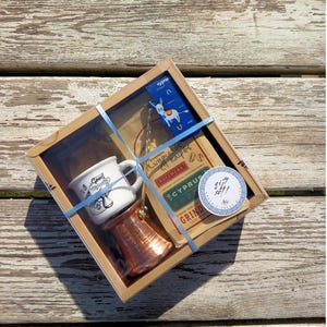 May include: A gift set for coffee lovers featuring a copper coffee pot, a white ceramic mug with the text "Good Morning" and a bag of Cyprus coffee. The set is packaged in a brown box with a blue ribbon and a sticker that says "Gift Set!!".