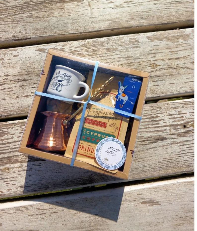 May include: A gift box set for making Greek coffee. The set includes a copper coffee pot, a small white mug, a coffee spoon, a bag of Cyprus coffee, and a blue and white sticker with a Greek key design.