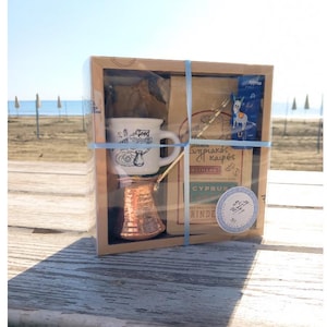 May include: A brown cardboard box with a clear plastic window, containing a small copper coffee pot, a white ceramic coffee cup with a blue and white design, and a bag of coffee. The box is tied with a blue ribbon and has a label that says "Greek Coffee Set".