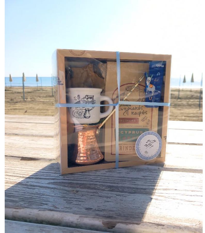 May include: A brown cardboard box with a clear plastic window, containing a small copper coffee pot, a white ceramic coffee cup with a blue and white design, and a bag of coffee. The box is tied with a blue ribbon and has a label that says "Greek Coffee Set".