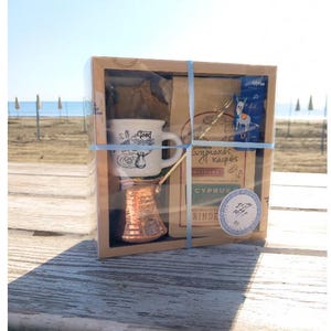 May include: A brown cardboard box with a clear plastic window, containing a small copper coffee pot, a white ceramic coffee cup with a blue and white design, and a bag of coffee. The box is tied with a blue ribbon and has a label that says "Greek Coffee Set".