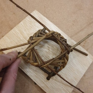 K&ouml;nnte beinhalten: Ein geflochtener Korb aus braunen Zweigen wird auf einer Holzbasis gebaut. Der Korb wird durch zwei lange Zweige gehalten, die sich kreuzen.