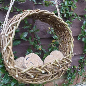 K&ouml;nnte beinhalten: Ein geflochtener Weidenvogel futterspender mit drei Talgkuchen drinnen. Der Futterspender h&auml;ngt an einem Seil und ist von gr&uuml;nem Laub umgeben.